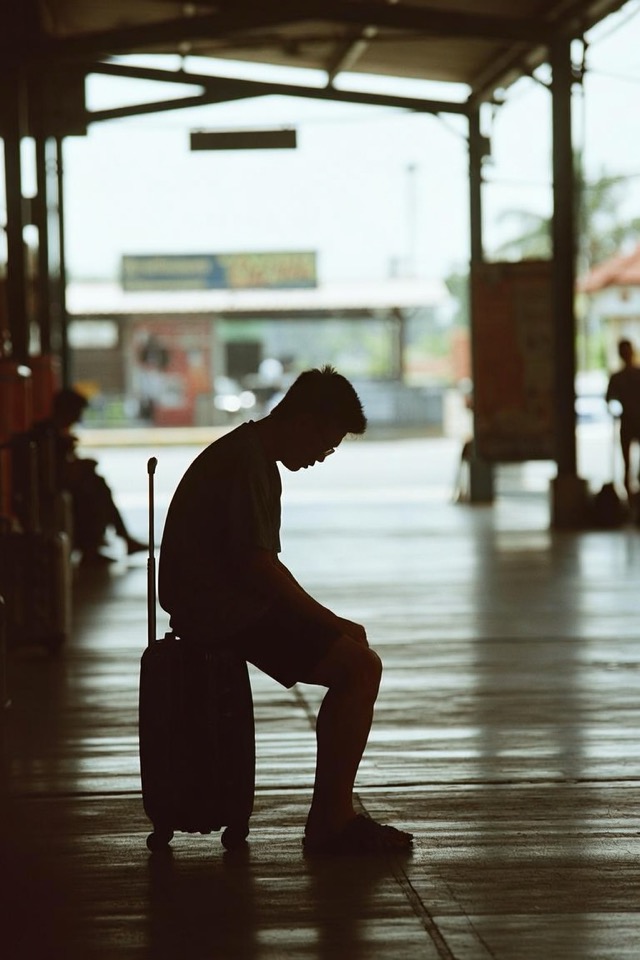 A tired traveler sitting on a rolling suitcase at a small provincial bus terminal
in Thailand, waiting under a covered outdoor platform