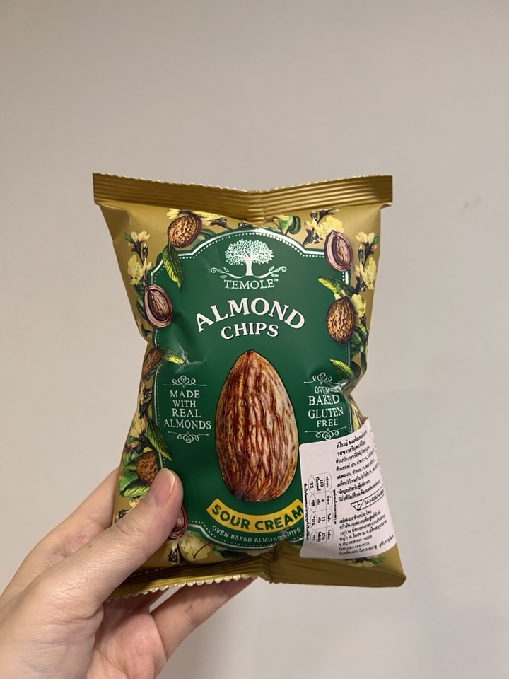 A bag of "Temole" brand Sour Cream Almond Chips being held by a hand. The bag is green and gold, with a central large almond, illustrative small almonds, and "Gluten Free" text. A label with Thai text and a barcode is attached to the bag
