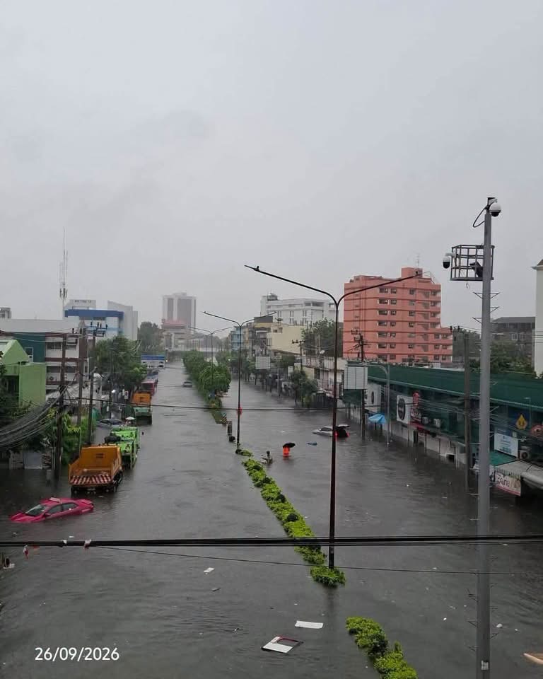 A panoramic view of a flooded city street on September 26, 2026. The street is submerged in water, and parked trucks and driving cars are visible.