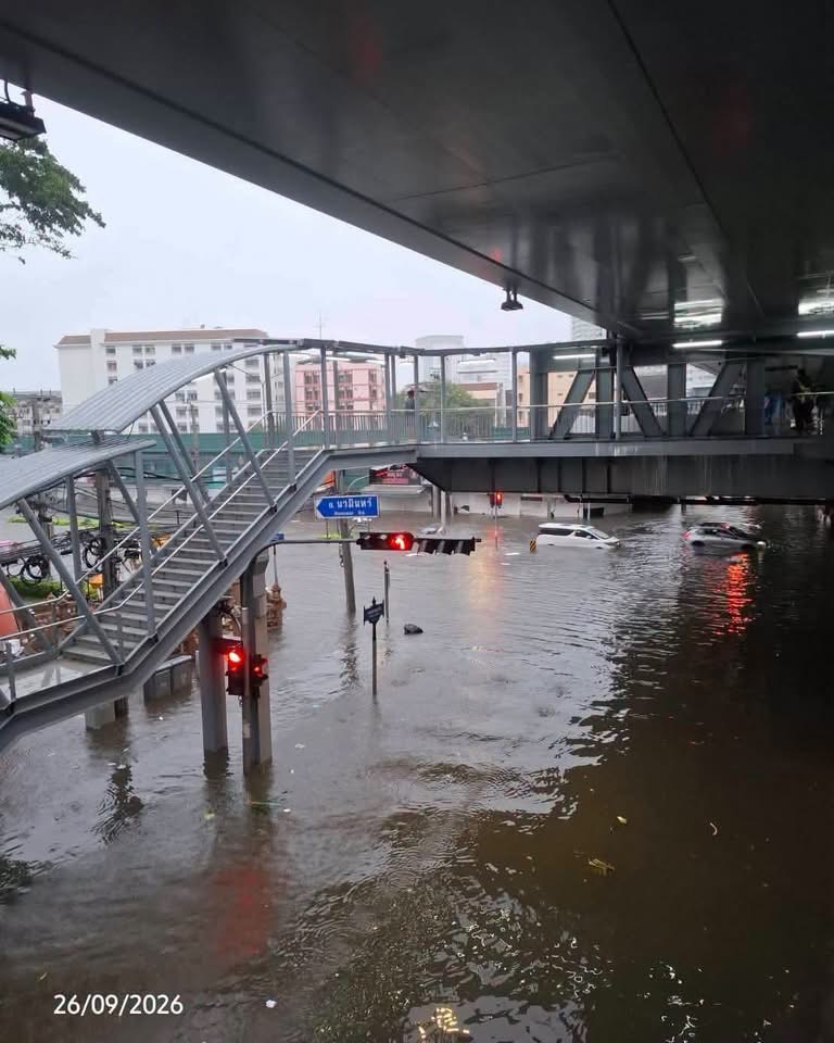A flooded city street on September 26, 2026. Water has flooded below an elevated pedestrian walkway and stairs, and cars are visible driving on the street.