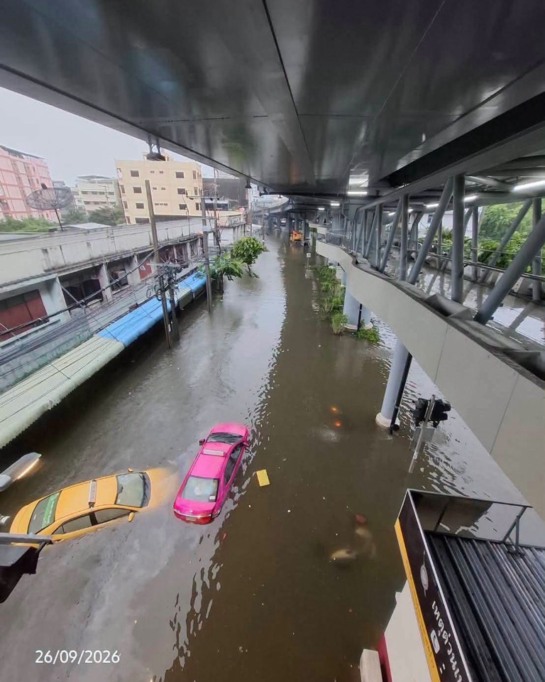 An overhead view of a flooded city street on September 26, 2026. A flooded street is visible under an elevated walkway, with pink and yellow cars floating on it.
