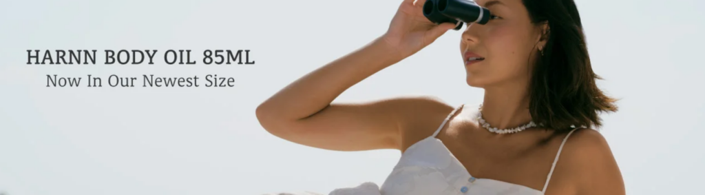 A woman holding binoculars while wearing a white dress and shell necklace, promoting HARNN Body Oil 85ml.